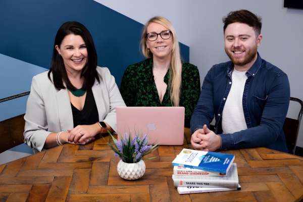 Three business colleagues sat around a table smiling at the camera: a white woman with dark hair, a white woman with blond hair (Samantha), and a man with dark hair. On the table are a pink laptop, plant and a pile of books. The background is blue an